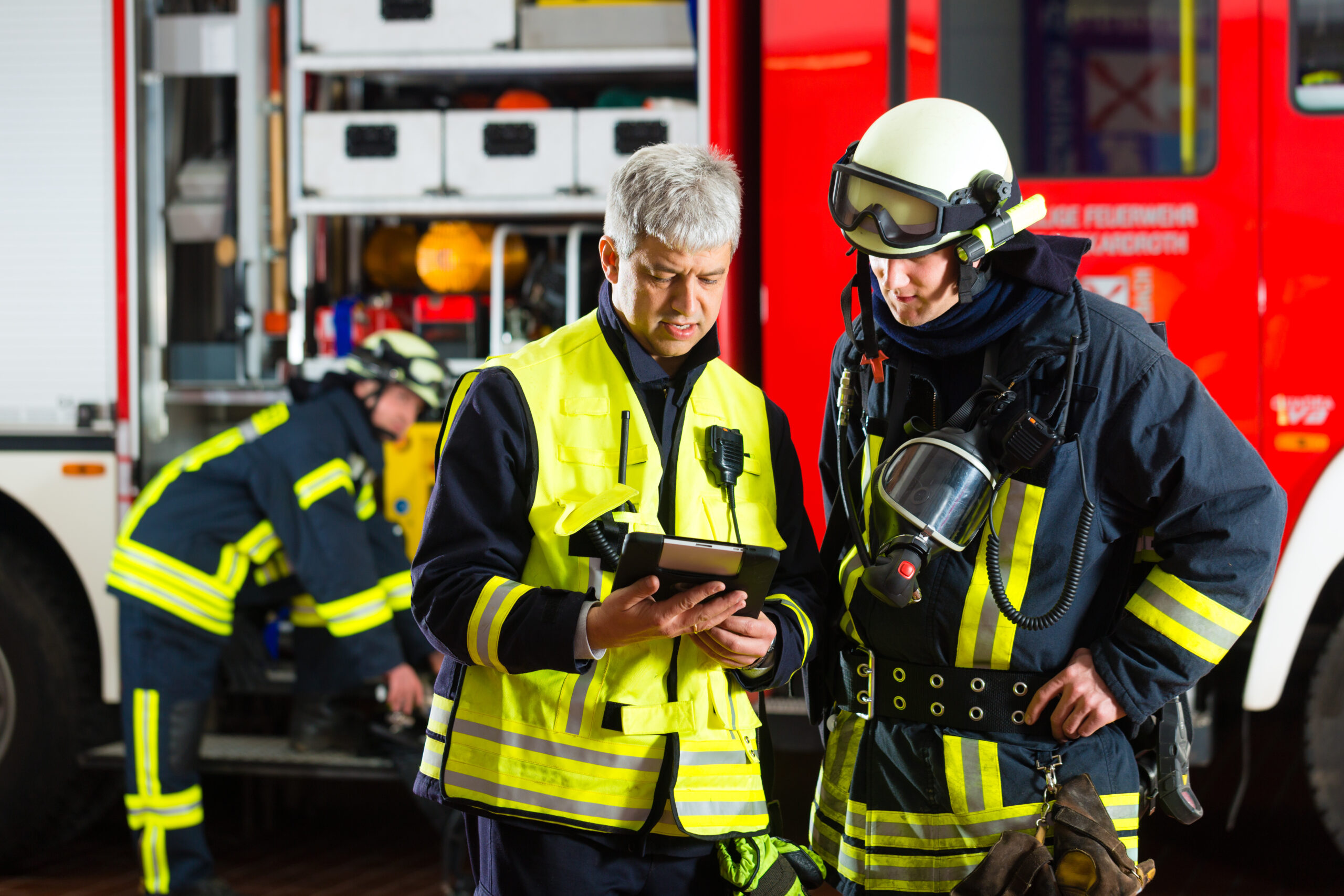 Einsatzleiter mit Tablet bespricht Ablauf mit Feuerwehrmann, im Hintergrund Ausrüstung und Löschfahrzeug.