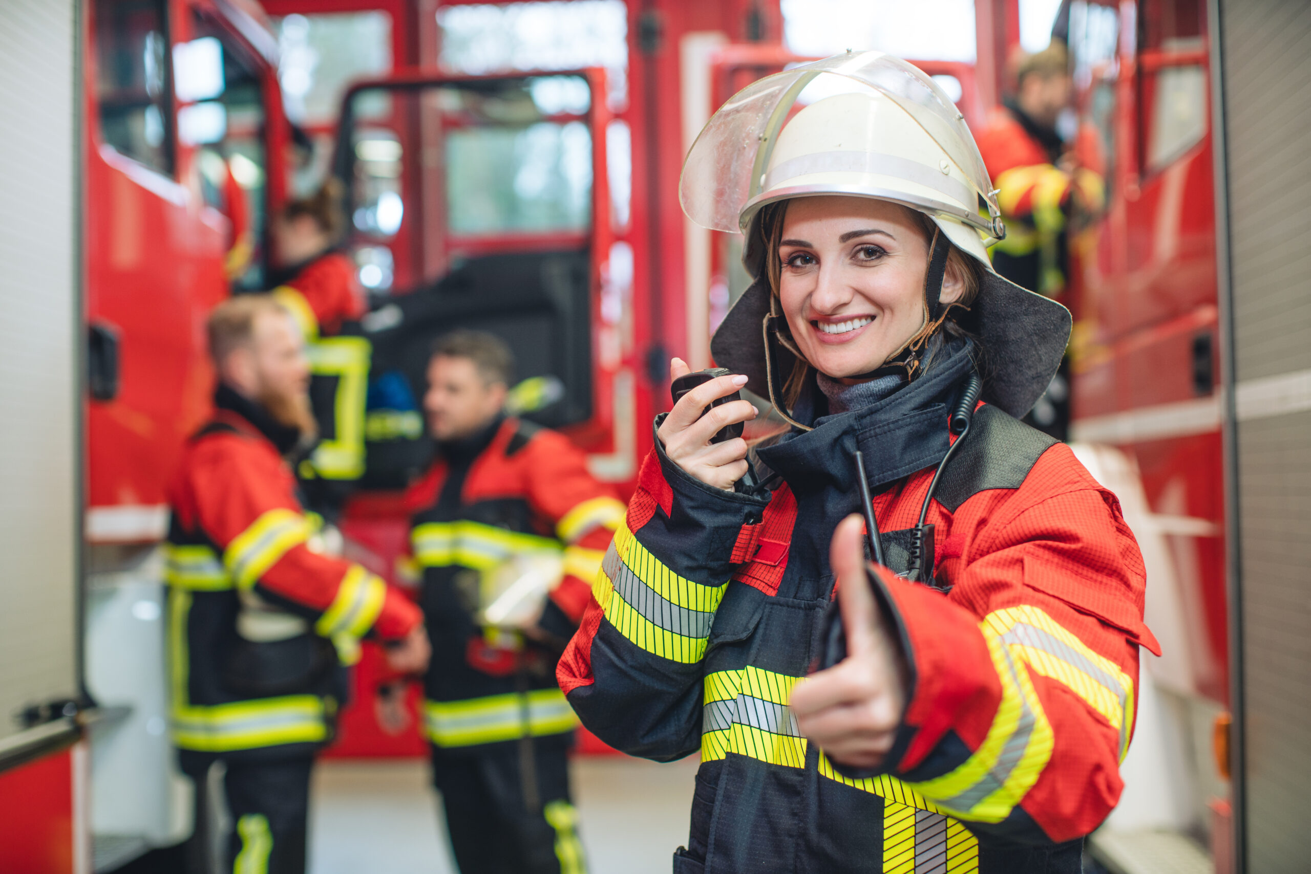 Lächelnde Feuerwehrfrau mit Funkgerät zeigt Daumen hoch, Team im Hintergrund am Löschfahrzeug.